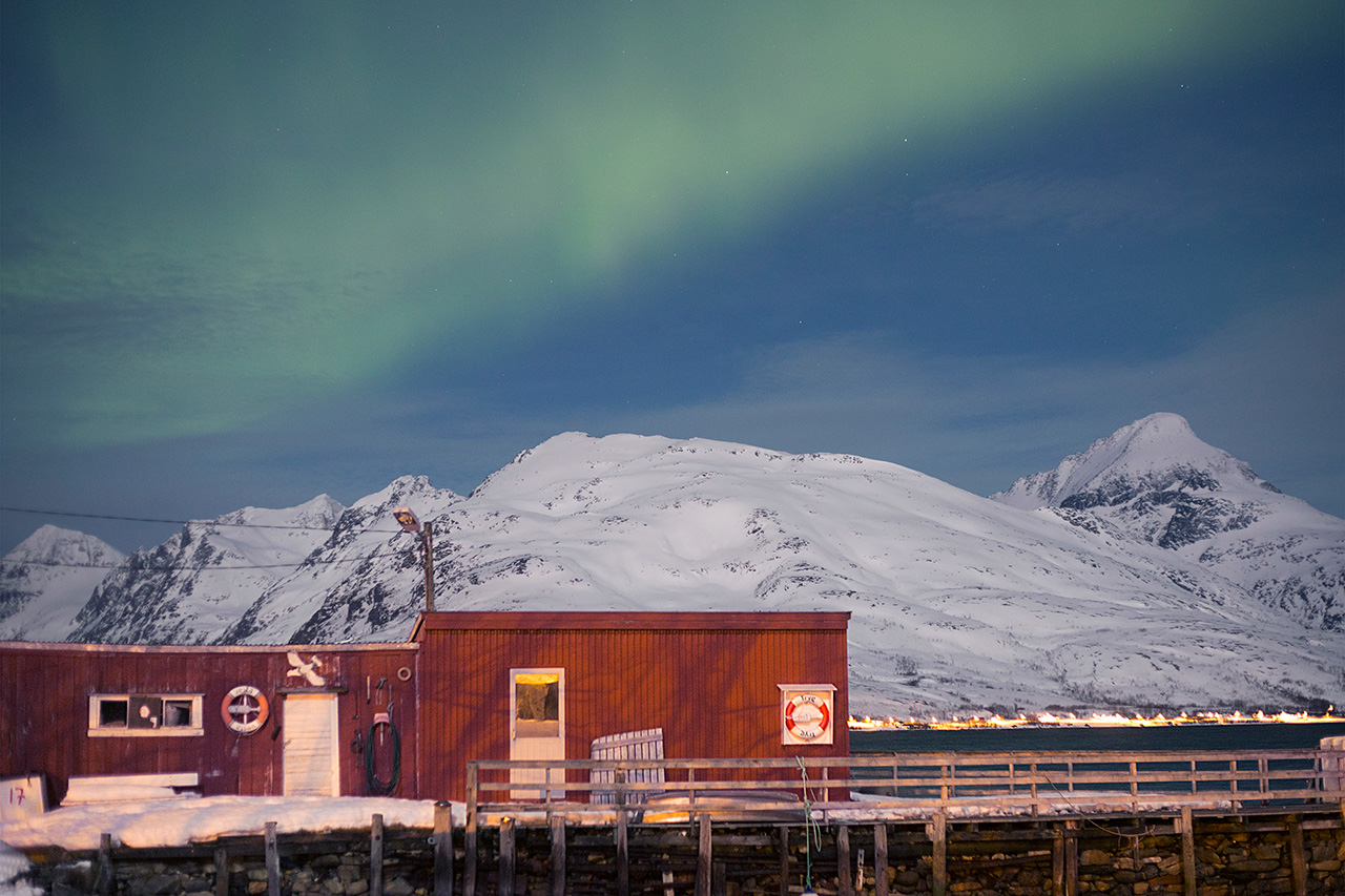 Northern lights over a red wooden boathouse and snowy mountains, Tromsø