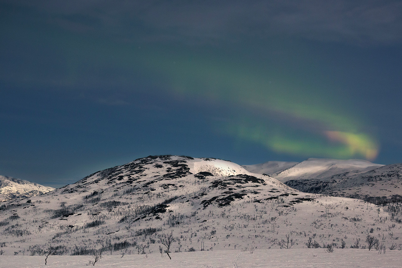 Aurora borealis arching across the sky above snow-covered hills, Tromsø