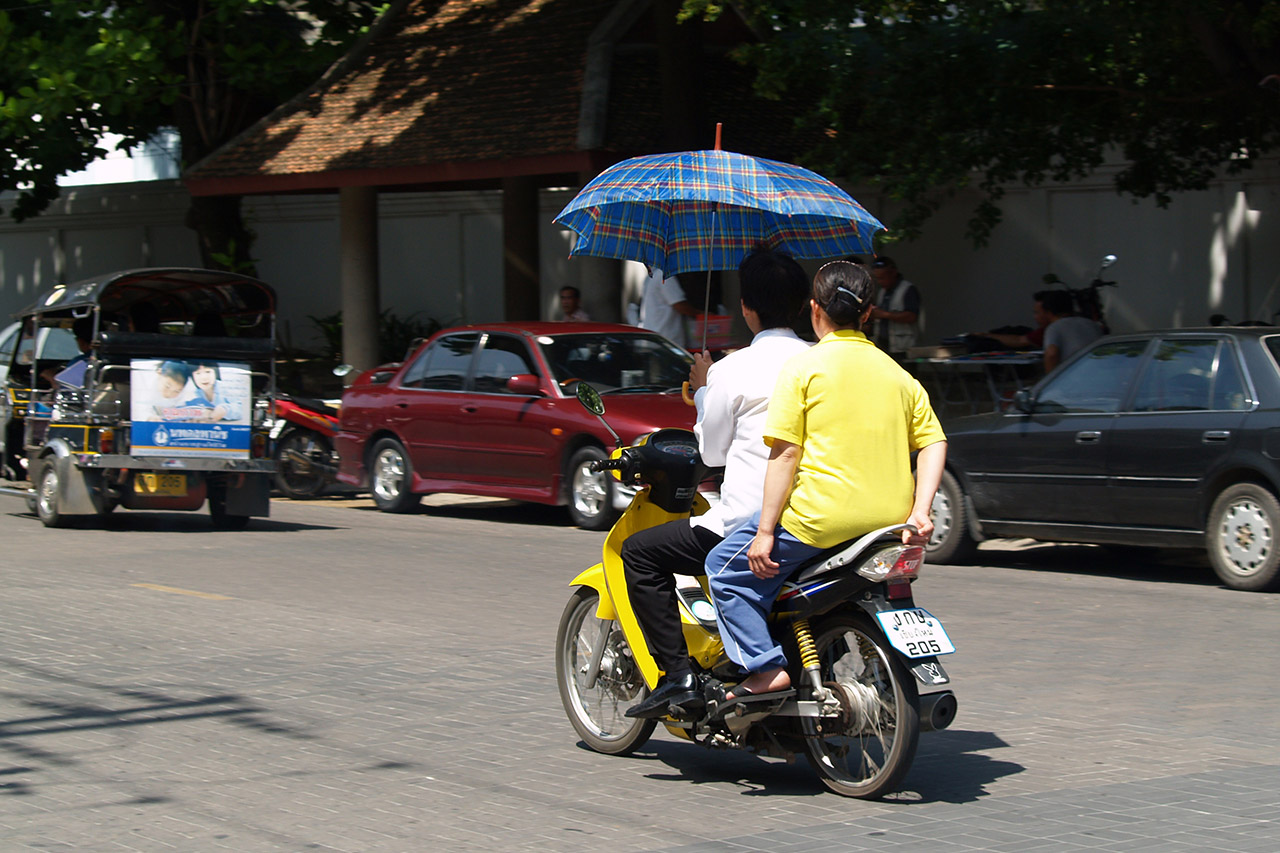 Two people on a yellow scooter under a blue umbrella in Bangkok traffic