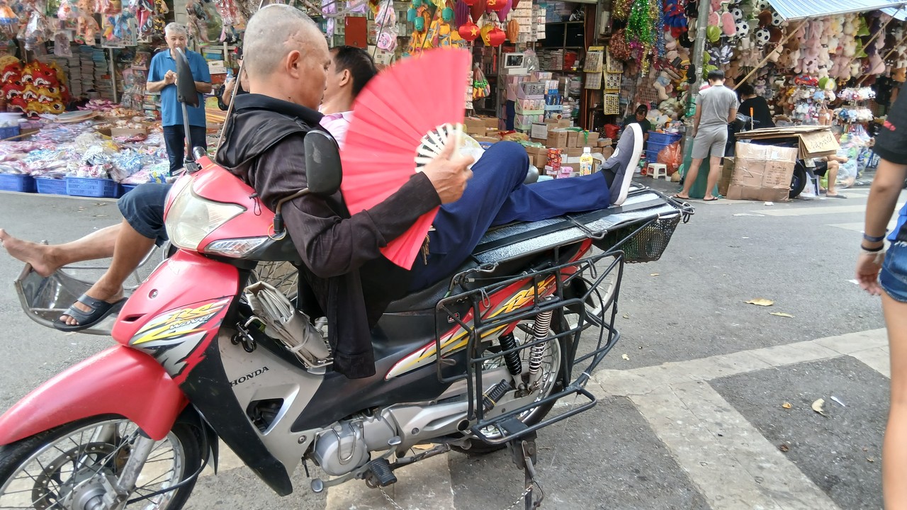 Men resting on a motorbike, one fanning himself with a red fan, in a Hanoi market street