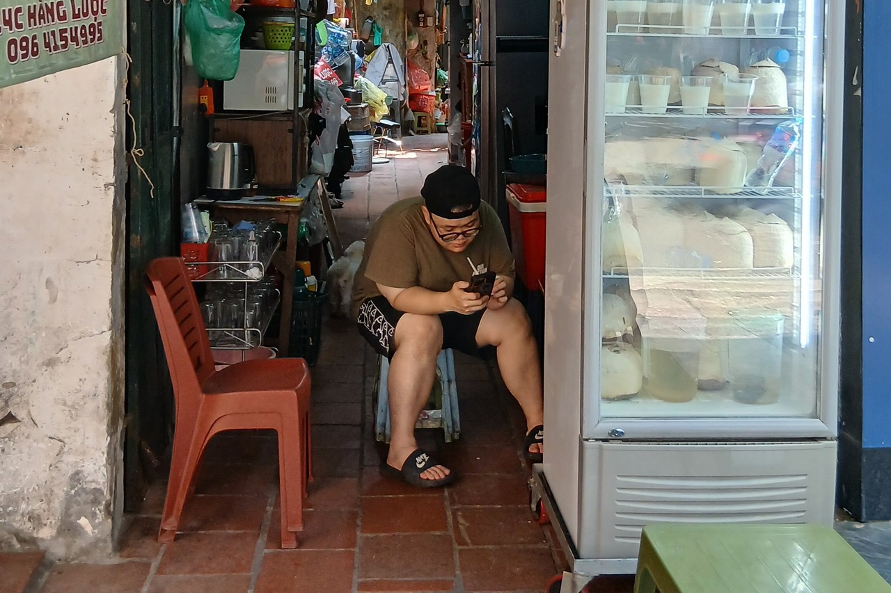 Man on a stool looking at his phone in a narrow Hanoi alleyway